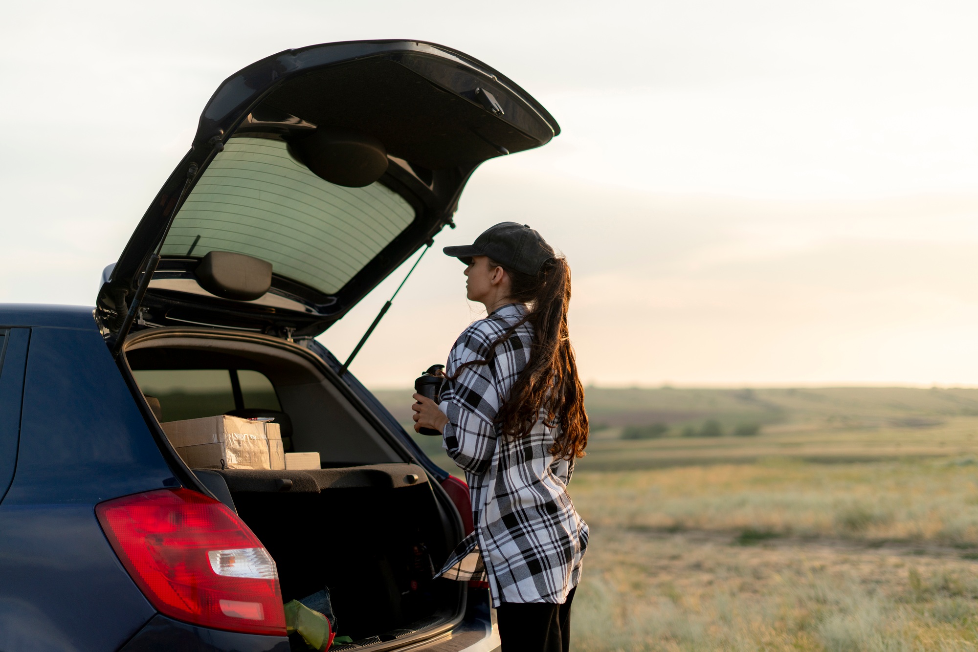 a young woman in a plaid shirt loads boxes into the trunk of her car, with the sunset